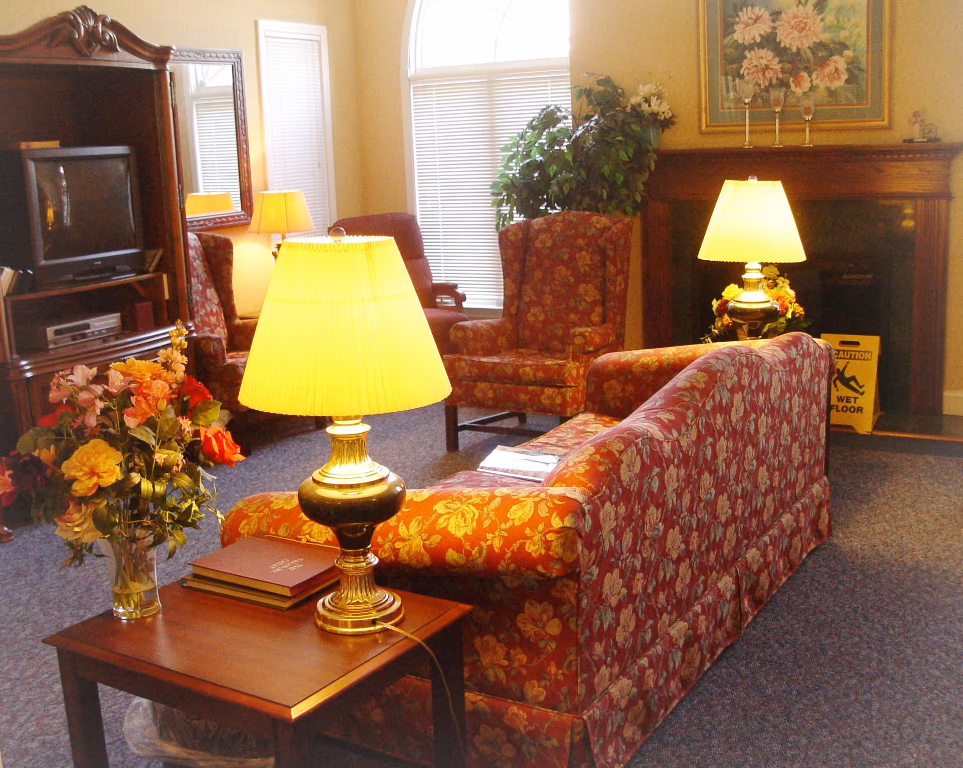 Cozy communal living room with floral-upholstered sofa and chairs, table lamps, TV and fireplace.