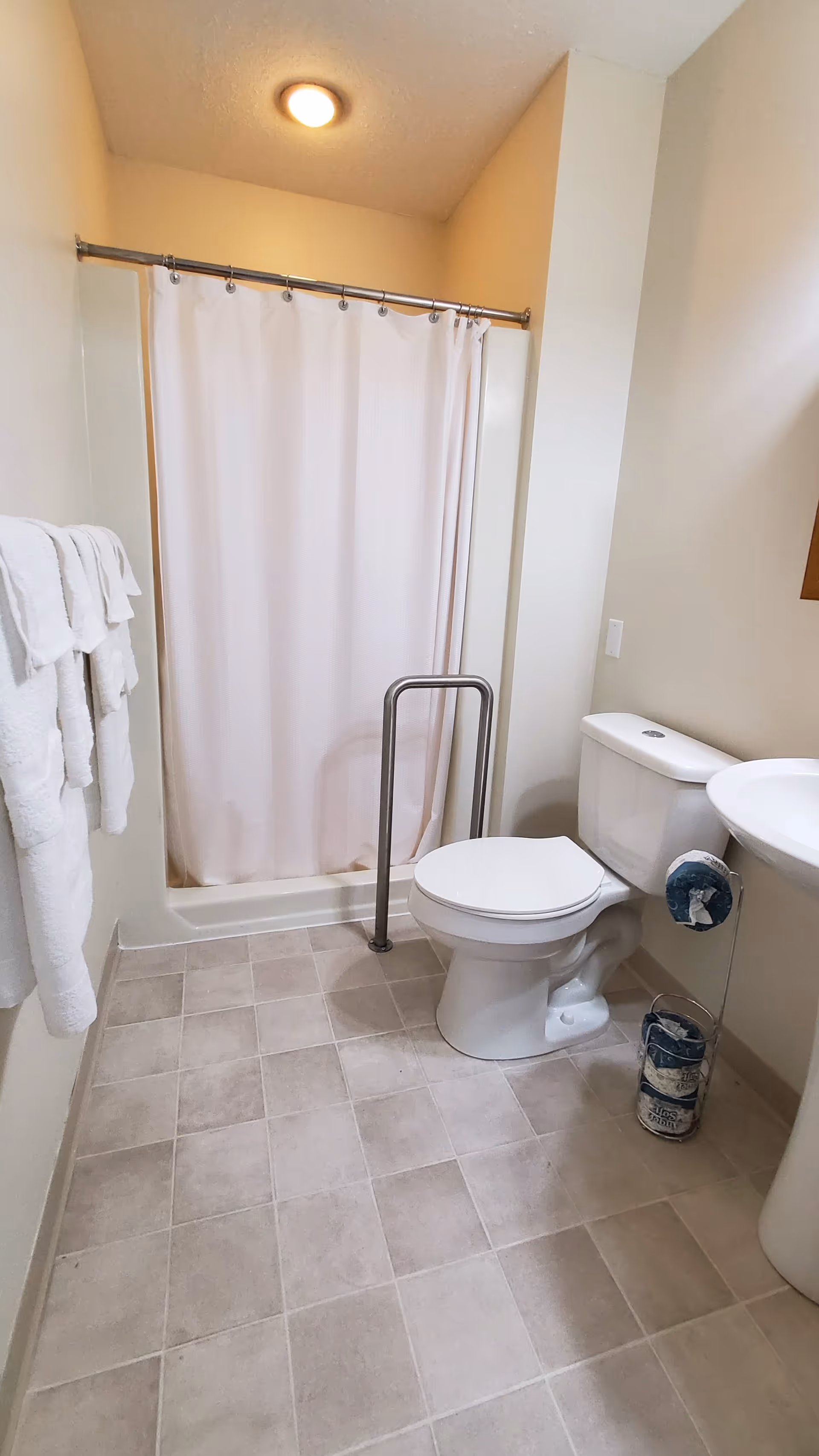 A clean bathroom with a shower area covered by a white curtain, a toilet with a support bar next to it, a pedestal sink, and several white towels hanging on a rack on the left wall. The floor is tiled with light-colored square tiles.