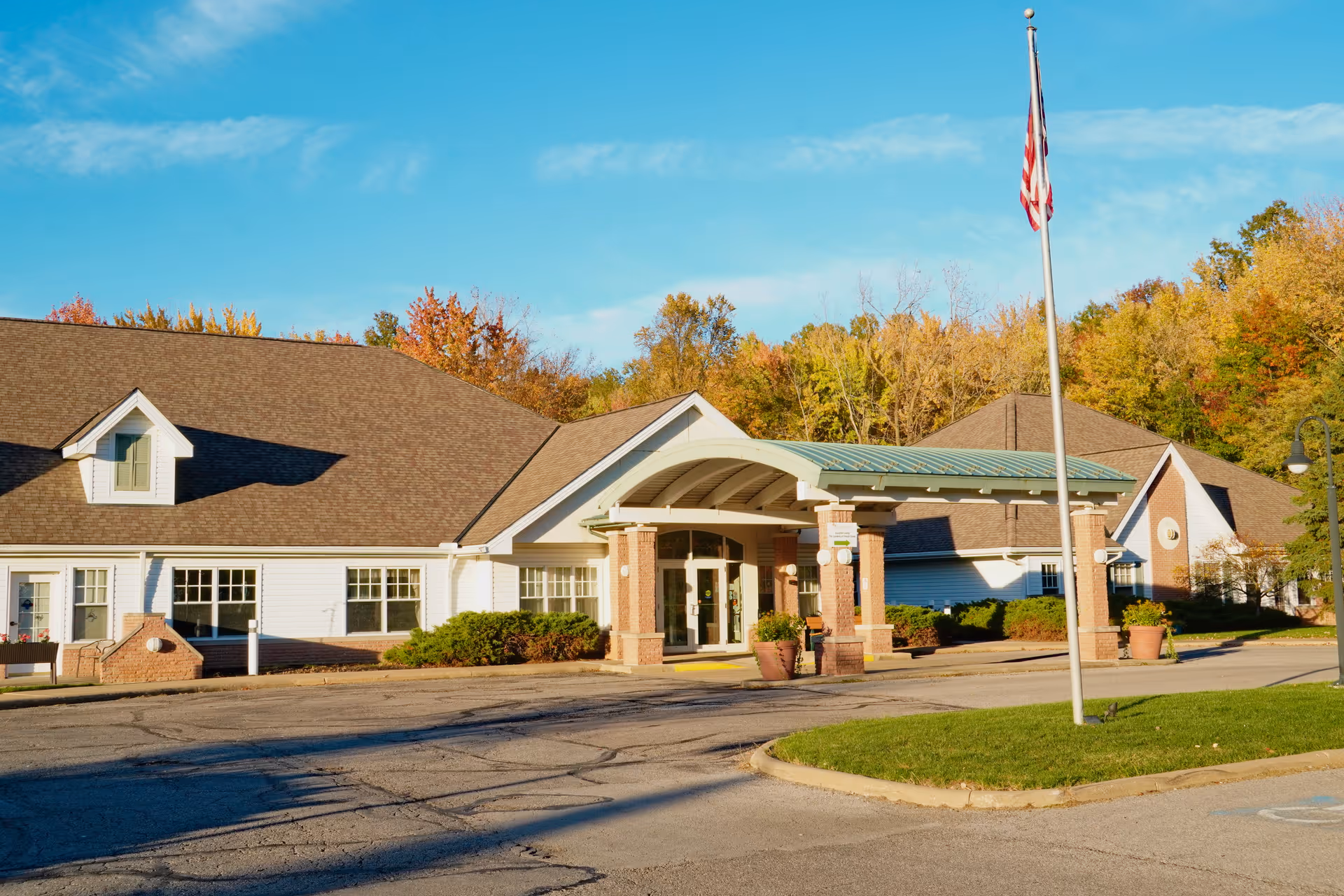 Exterior view of Avon Oaks Caring Community building with a covered entrance, an American flag on a flagpole, and trees with autumn foliage in the background under a clear blue sky.