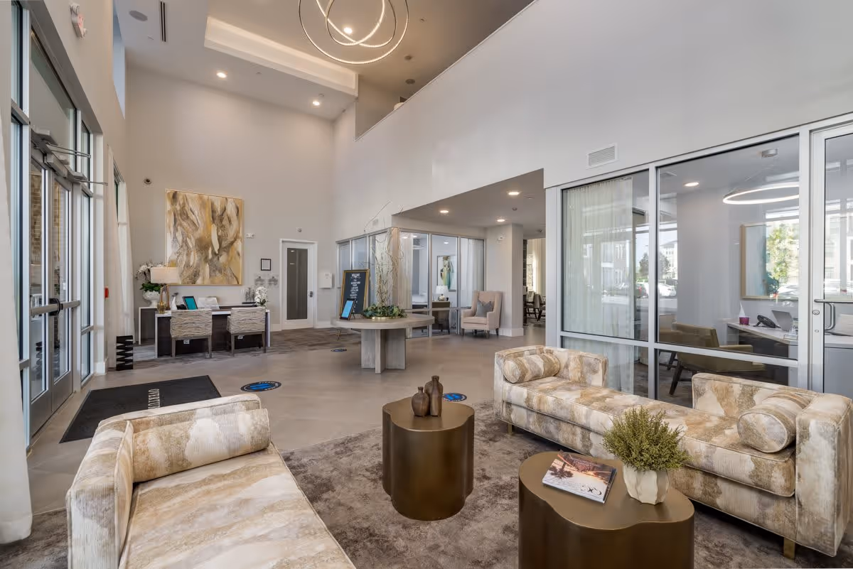 A modern, spacious senior living facility lobby with high ceilings, two beige patterned sofas facing each other, two round bronze coffee tables with decorative items, a round table with a plant centerpiece, and a reception desk with two chairs. Large windows and glass doors allow natural light to fill the space, and there is abstract artwork on the wall behind the reception desk.