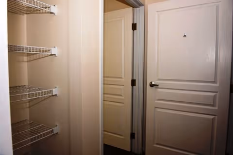 Interior view of a hallway with beige walls and two closed white doors, one on the right and one straight ahead. On the left side, there are white wire shelves mounted on the wall.