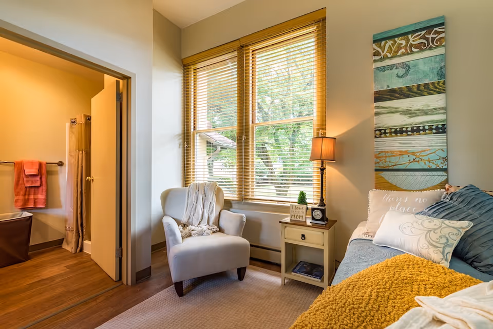 A cozy room with a large window featuring wooden blinds, a comfortable armchair with a throw blanket, a small side table with a lamp and decorative items, and a daybed with multiple pillows and a yellow knitted blanket. An open door reveals a bathroom with a shower curtain and towels hanging on a rack.