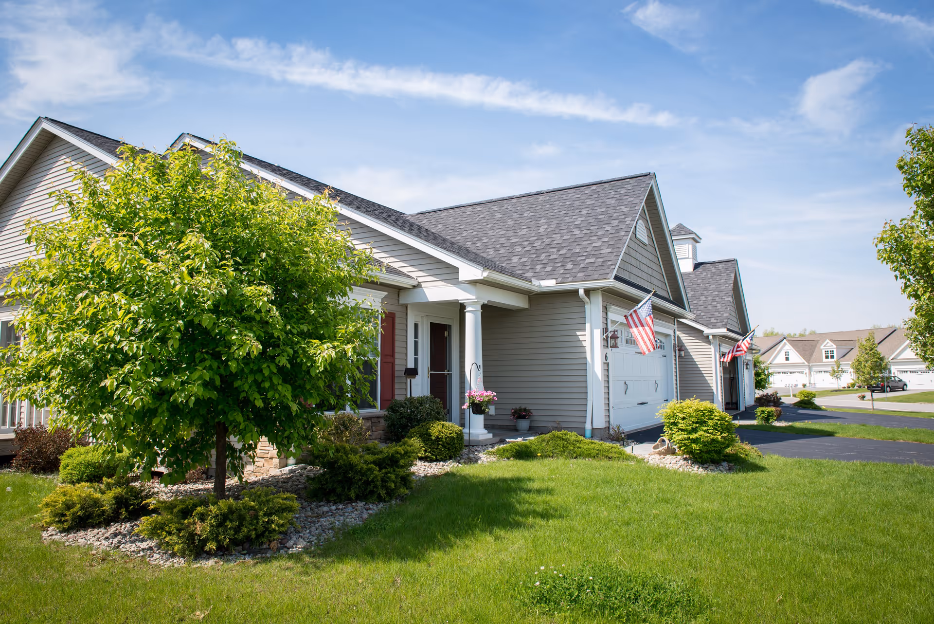 Exterior view of a residential building with beige siding, a dark shingled roof, and a white garage door. The front yard has green grass, bushes, and a tree. Two American flags are displayed near the garage. The sky is blue with some clouds.