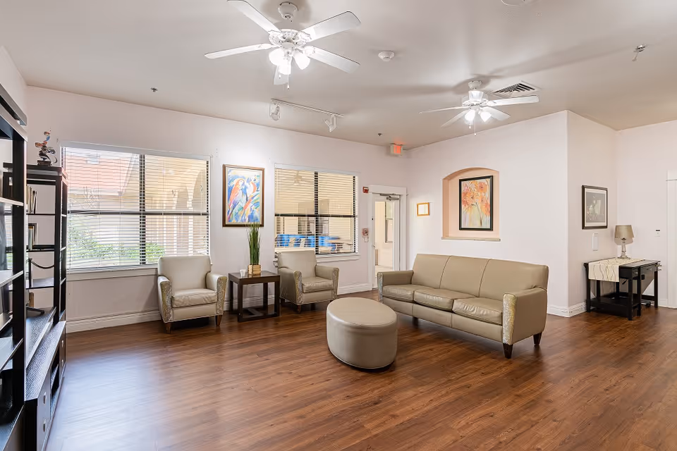 A bright and spacious living room area in an assisted living facility featuring a beige leather sofa, two matching armchairs, a round ottoman, wooden flooring, ceiling fans with lights, large windows with blinds, colorful framed artwork on the walls, and a black bookshelf on the left side.
