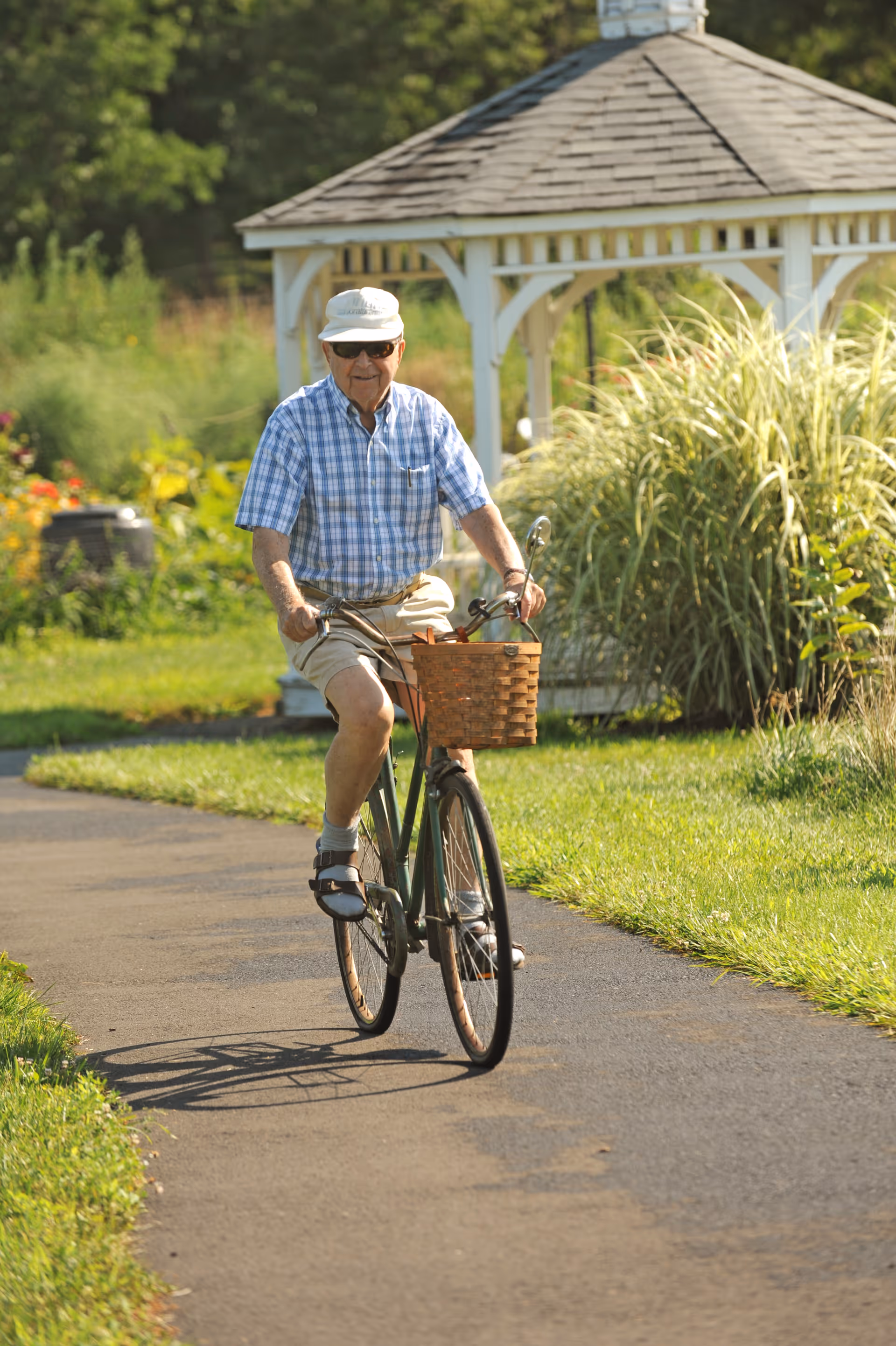 An elderly man wearing a white hat, sunglasses, a blue checkered shirt, and beige shorts is riding a bicycle with a wicker basket on a paved path. Behind him is a white gazebo surrounded by greenery and tall grass on a sunny day.
