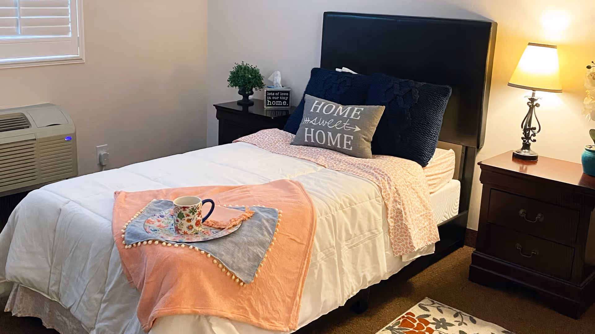 A cozy bedroom with a single bed featuring a black headboard, white bedding, a pink blanket, and decorative pillows including one that says 'HOME sweet HOME'. There is a wooden nightstand on each side of the bed, one with a lamp and a small plant, and the other with a small plant and a tissue box. A colorful floral rug is partially visible on the floor.
