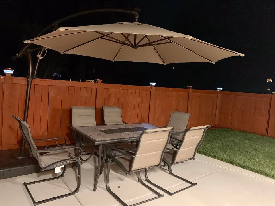 Outdoor patio area at night with a large beige umbrella over a rectangular table surrounded by six beige and black metal chairs, next to a wooden fence and a patch of green grass.