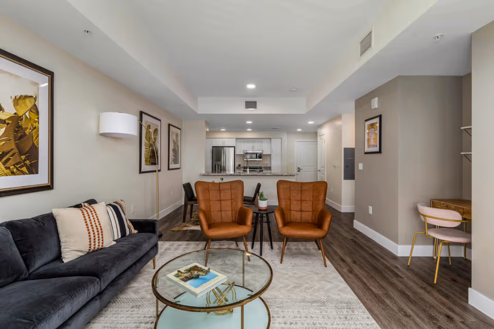 A modern living room with a dark gray sofa, two brown leather armchairs, a glass coffee table with books, and framed artwork on the walls. The room has wood flooring and leads into a kitchen area with white cabinets and stainless steel appliances. There is also a small desk with a pink chair in the corner.