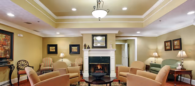 A cozy living room with a lit fireplace centered on the far wall, surrounded by various upholstered chairs and a green sofa. The room features warm yellow walls, framed artwork, table lamps on side tables, and a ceiling light fixture with recessed lighting. A round coffee table is placed in front of the fireplace.