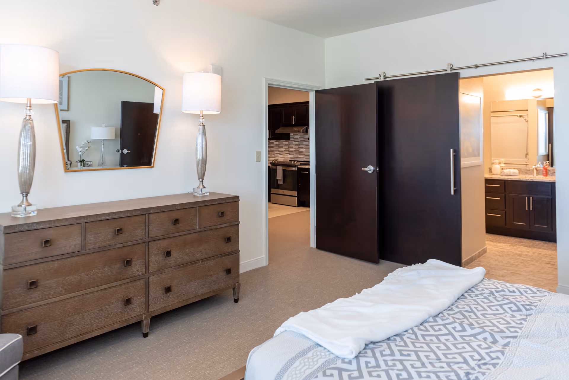 A bedroom with a wooden dresser topped with two tall lamps and a mirror above it. The bed is partially visible with a patterned blanket and a white throw. There are two dark wooden doors, one leading to a kitchen and the other to a bathroom with a sink and mirror visible.