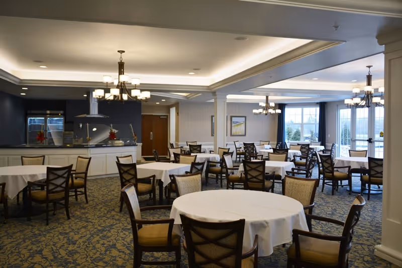 Spacious dining room with round tables covered in white tablecloths, multiple chairs, chandeliers, and large windows.
