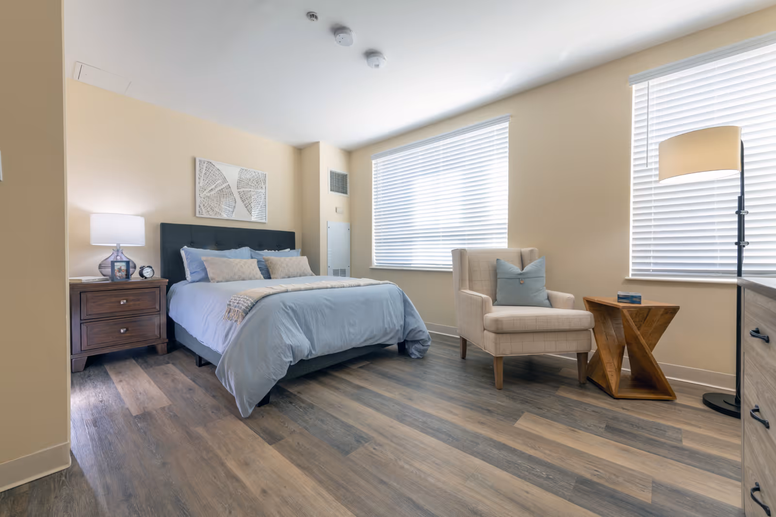 Well-lit bedroom with a neatly made bed, nightstand and lamp, an upholstered armchair and side table by large windows.