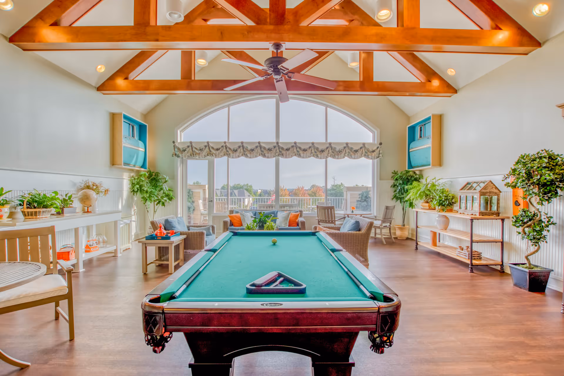 A bright and spacious recreational room with a pool table in the center, surrounded by comfortable seating including armchairs and small tables. The room features large arched windows letting in natural light, wooden ceiling beams, a ceiling fan, and various plants and decorative items along the walls.