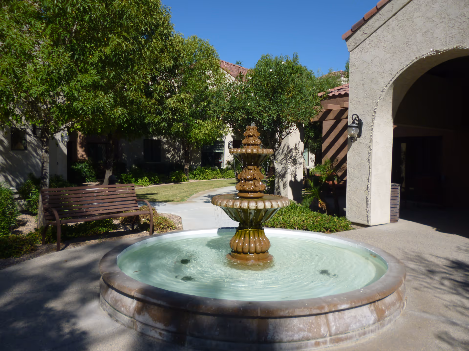A sunny courtyard showing a three-tier stone fountain surrounded by a bench, trees, and the stucco entrance to a building.