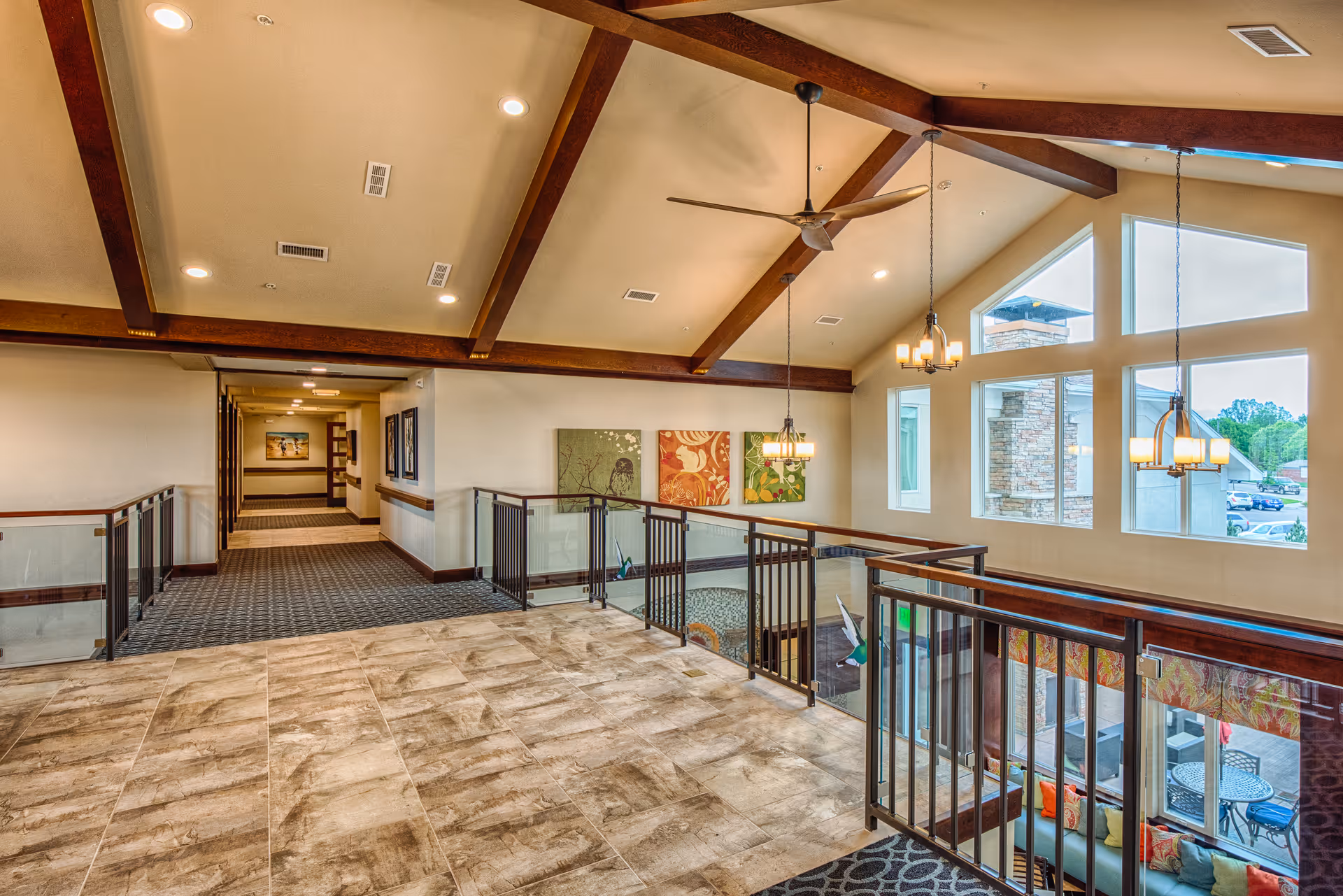 Interior view of a senior living facility hallway with a high ceiling featuring wooden beams and ceiling fans. The area has large windows allowing natural light to enter, modern hanging light fixtures, and a railing overlooking a lower level with seating and a patio area outside. The floor is tiled near the railing and carpeted in the hallway, with artwork on the walls.