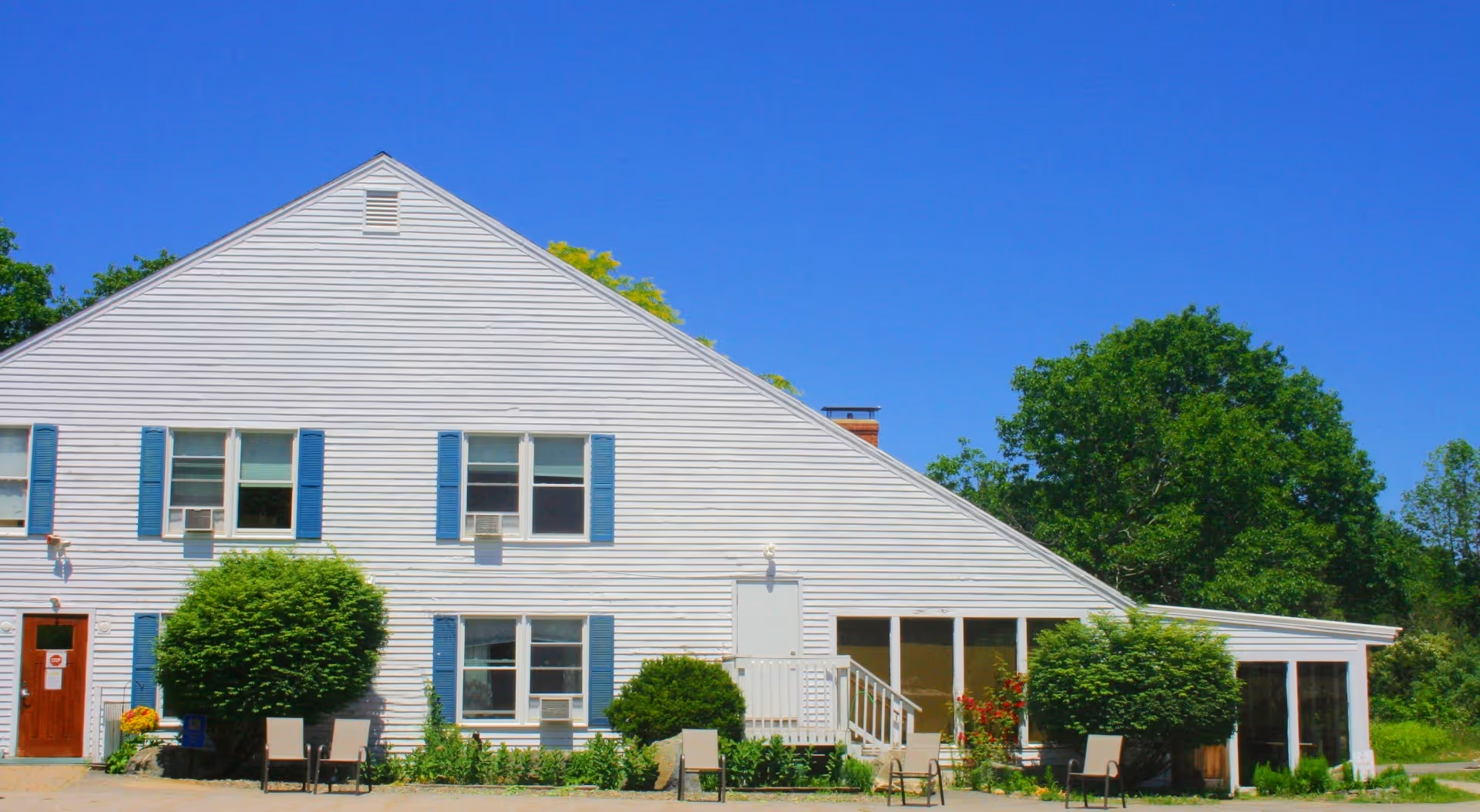 White two-story residential building with blue shutters, a small porch, chairs and trimmed shrubs under a clear blue sky.