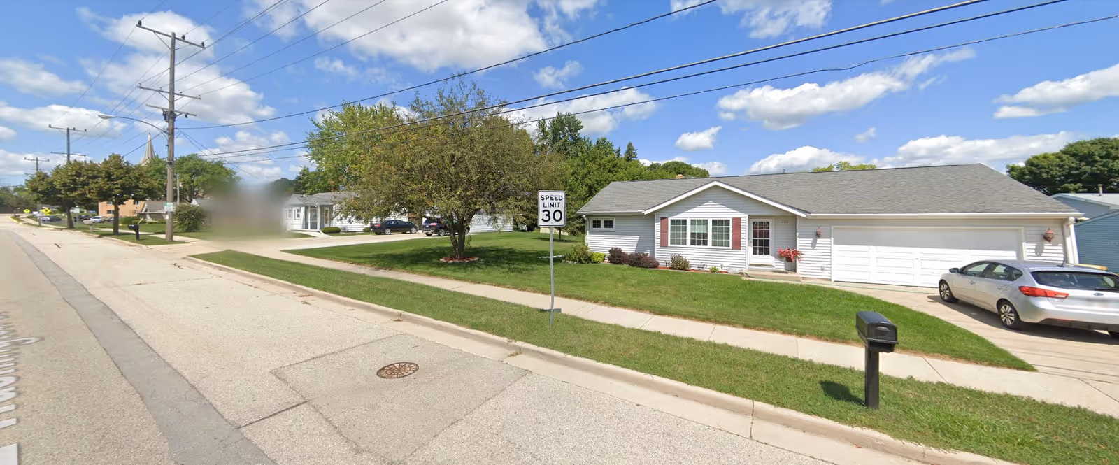 Street view of a residential neighborhood with a single-story house featuring a gray roof, white siding, and a two-car garage. A car is parked in the driveway, and there is a mailbox on the grass near the sidewalk. A speed limit sign indicating 30 mph is visible on the lawn. Trees and other houses line the street under a partly cloudy sky.