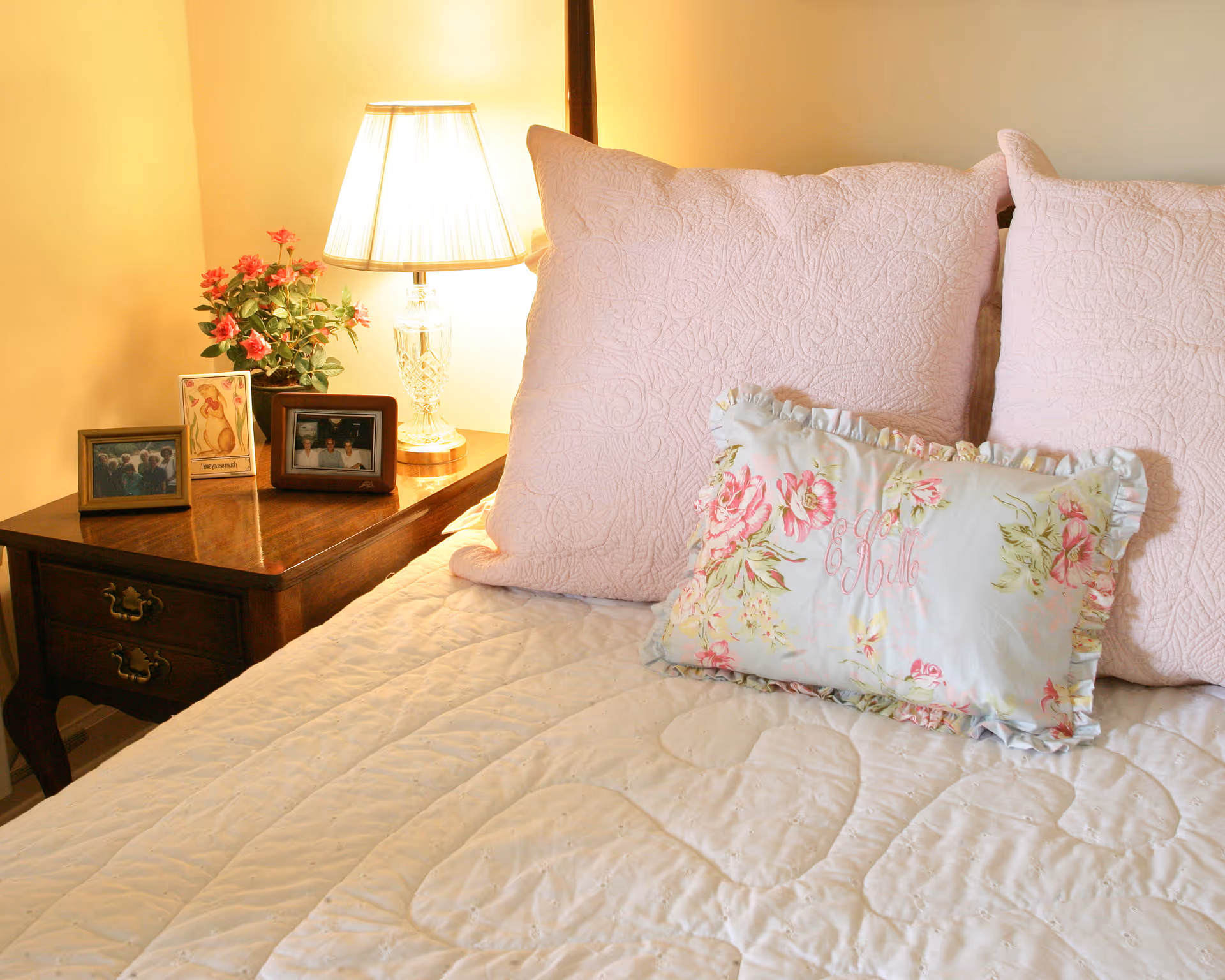 A cozy bedroom corner featuring a bed with a white quilted bedspread, two large pink pillows, and a smaller floral decorative pillow. Next to the bed is a wooden nightstand with two drawers, holding a crystal table lamp with a white shade, a small potted plant with pink flowers, and three framed photographs.