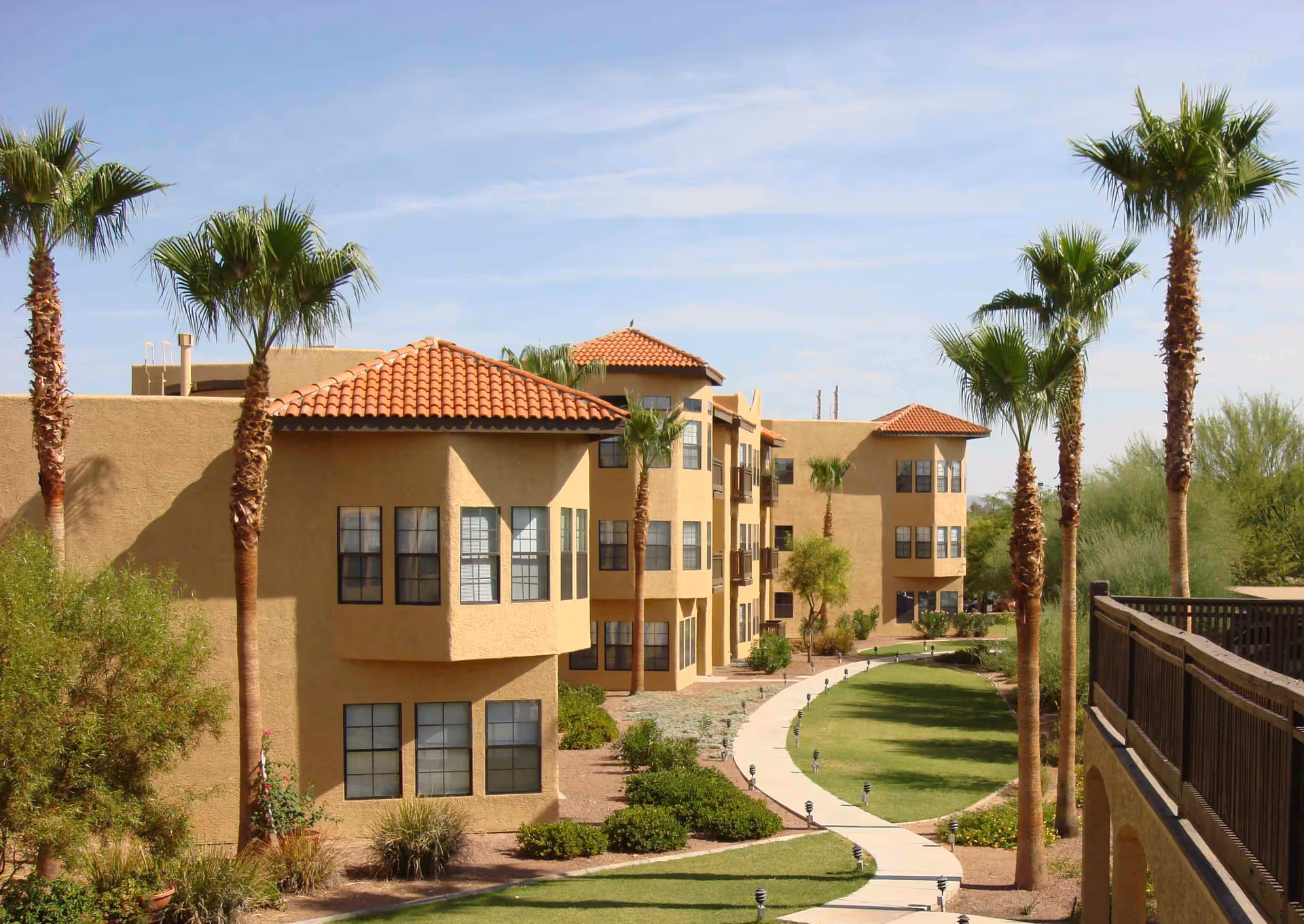 Exterior view of a senior living facility with beige stucco buildings featuring red tile roofs, surrounded by palm trees and landscaped greenery. A curved concrete walkway runs through a grassy area between the buildings and a wooden railing on the right side.