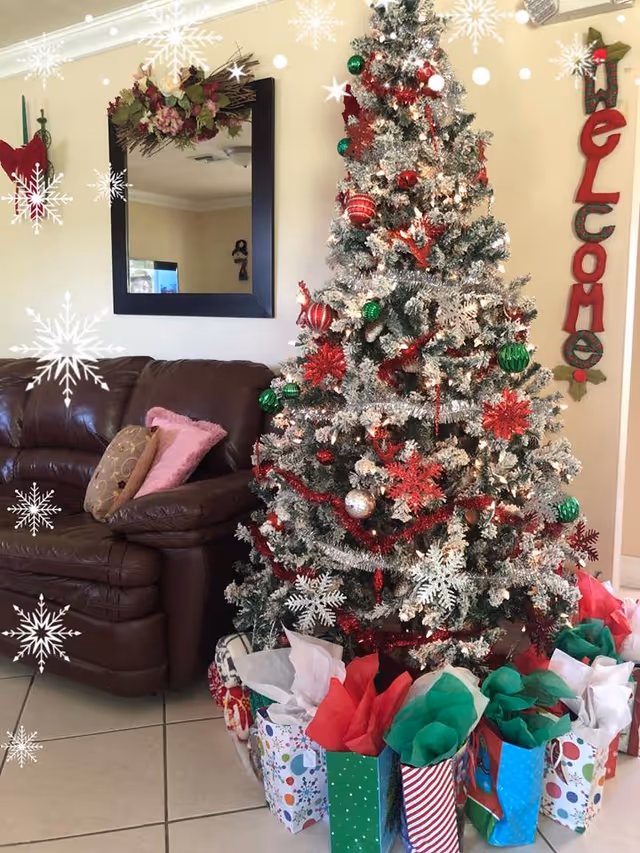A decorated Christmas tree with red, green, and silver ornaments and garlands stands next to a brown leather couch with pillows. Several colorful gift bags with tissue paper are placed under the tree. A large mirror with floral decoration hangs on the wall above the couch, and a vertical 'Welcome' sign is mounted on the wall beside the tree. Snowflake graphics overlay the image.