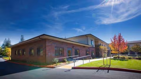 Brick, one-story senior living facility exterior with ramp, manicured lawn, and blue sky.
