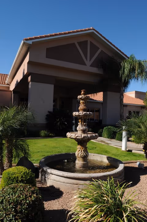 Outdoor view of a senior living community entrance featuring a multi-tiered stone fountain surrounded by green shrubs and palm trees, with a building having a tiled roof and covered entrance in the background under a clear blue sky.