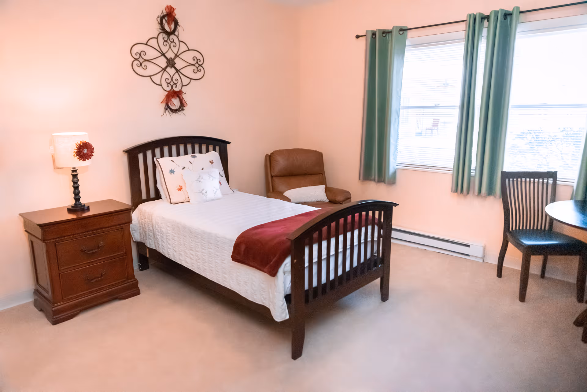 A tidy single bedroom with a wooden bed, nightstand and lamp, armchair, wall decor, and a window with green curtains.