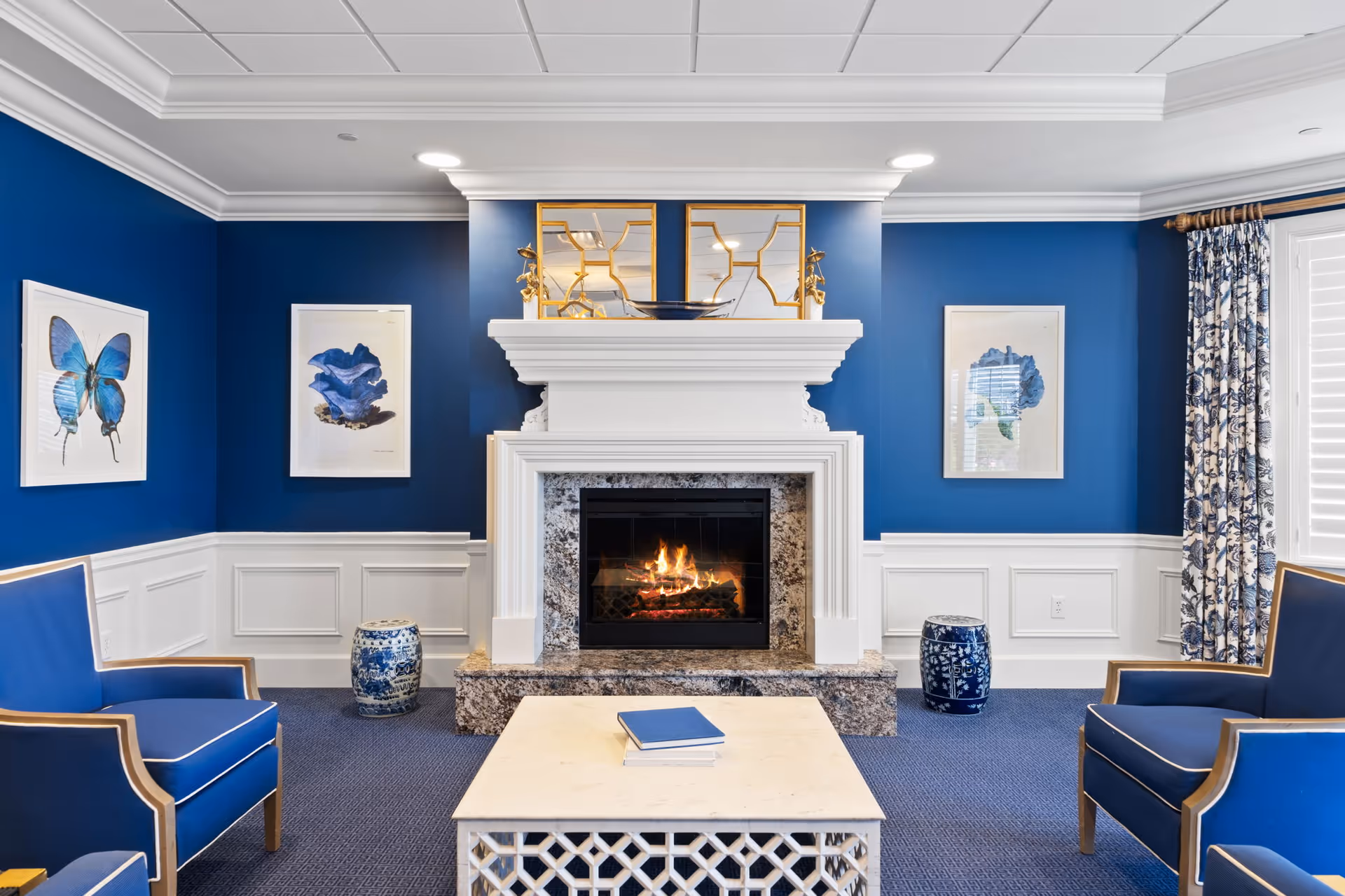 A cozy living room with blue walls and white wainscoting, featuring a white fireplace with a burning fire. Above the fireplace are two decorative mirrors and a bowl. On either side of the fireplace are framed blue-themed artworks. Two blue armchairs with wooden frames face a white coffee table with a book on it. Blue patterned curtains hang by a window with white shutters.
