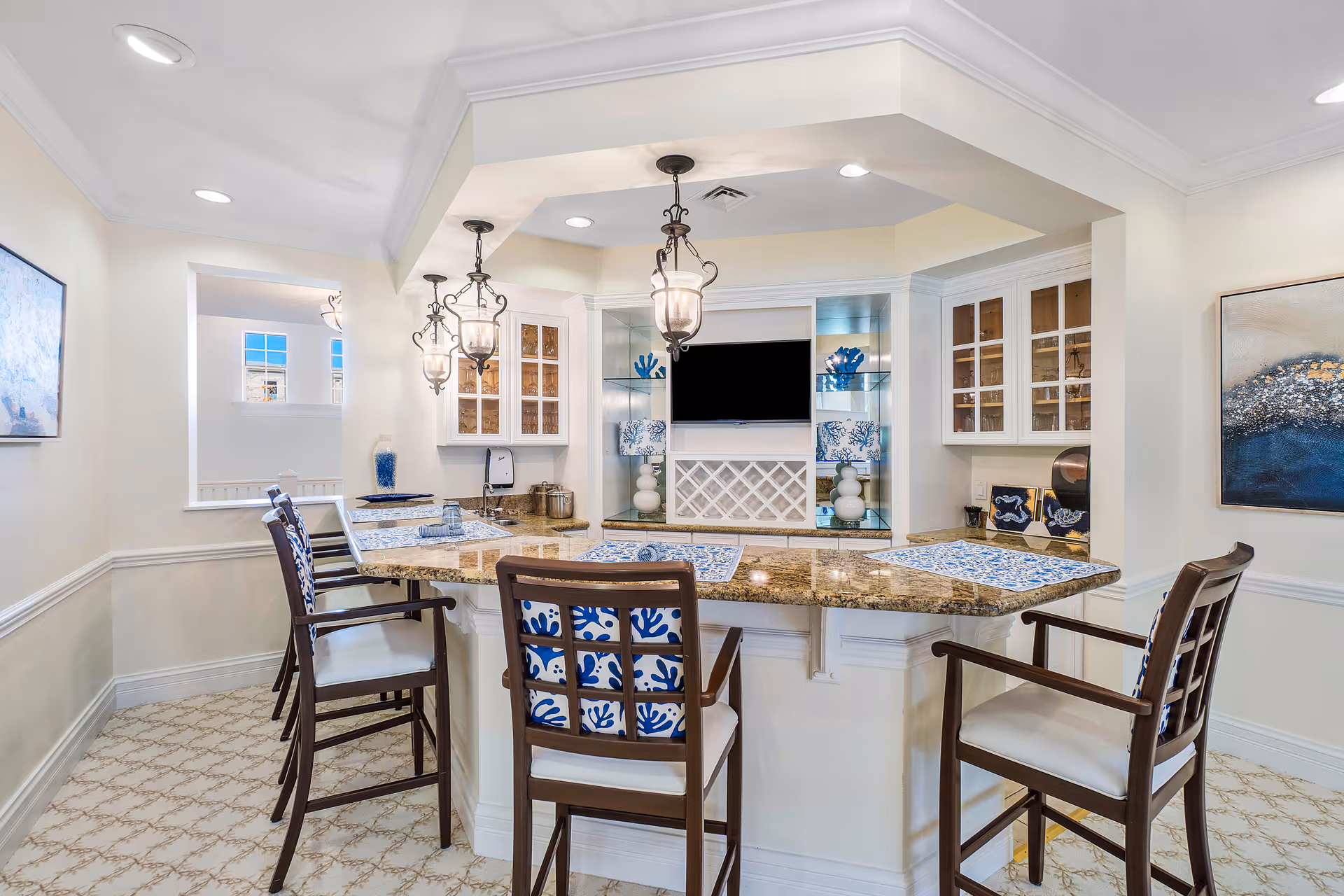 Bright kitchenette/bar area with a granite counter, four bar stools, pendant lights, glass-front cabinets and a small TV.