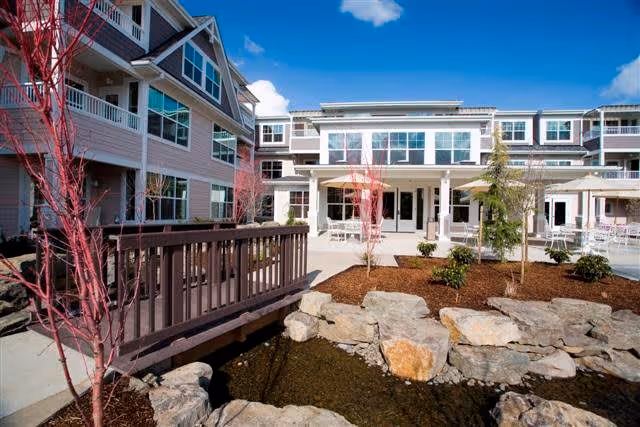 Outdoor courtyard area of The Stafford Retirement Community featuring a small wooden bridge over a rock-lined pond, landscaped garden beds with young trees and shrubs, and a multi-story building with large windows and balconies under a bright blue sky with some clouds.
