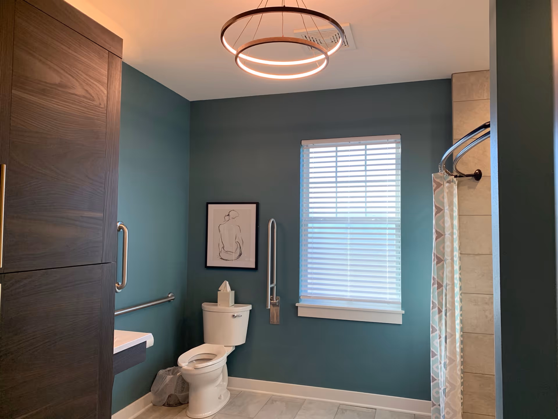 A bathroom with teal walls featuring a white toilet with a tissue box on top, a window with white blinds, a framed minimalist line drawing of a seated person, a grab bar next to the toilet, a dark wood cabinet, a modern circular ceiling light, and a shower with a patterned curtain on the right side.