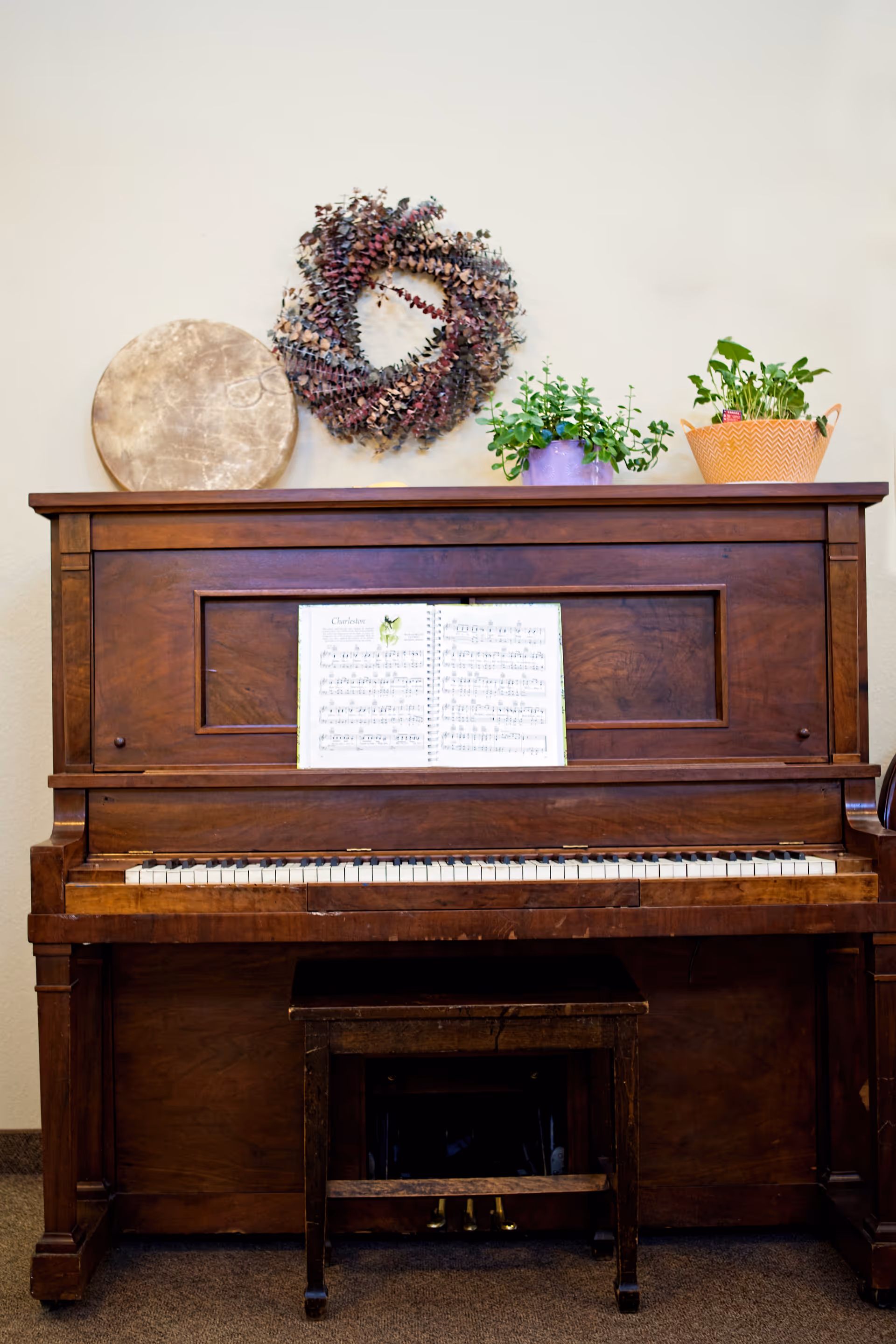 A wooden upright piano with an open sheet of music on the music stand. On top of the piano are a round drum, a decorative wreath, and two potted plants. A small wooden bench is positioned in front of the piano.
