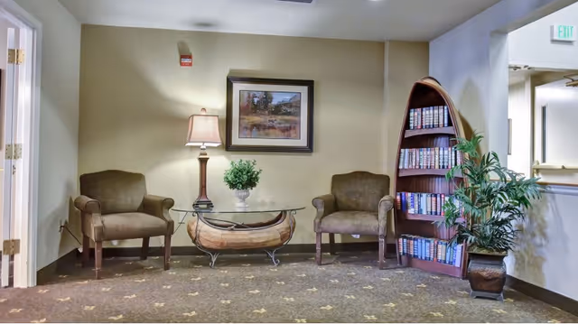 Small lounge area with two upholstered armchairs, a glass-top coffee table, a table lamp and framed artwork, a boat-shaped bookcase of books, and a potted plant.