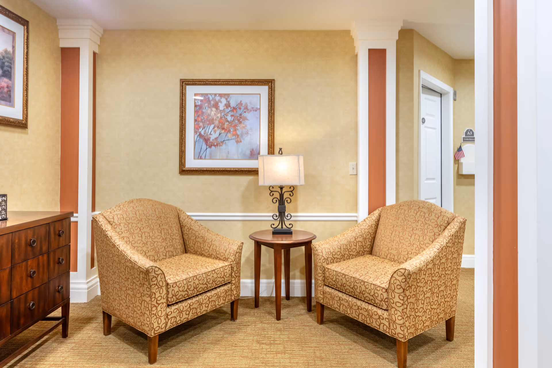 A cozy sitting area with two patterned armchairs facing each other with a small round wooden table and a decorative lamp between them. The walls are beige with white trim and a framed picture of autumn leaves hangs above the table. To the left is a wooden dresser, and to the right is a hallway with a white door and an American flag decoration.