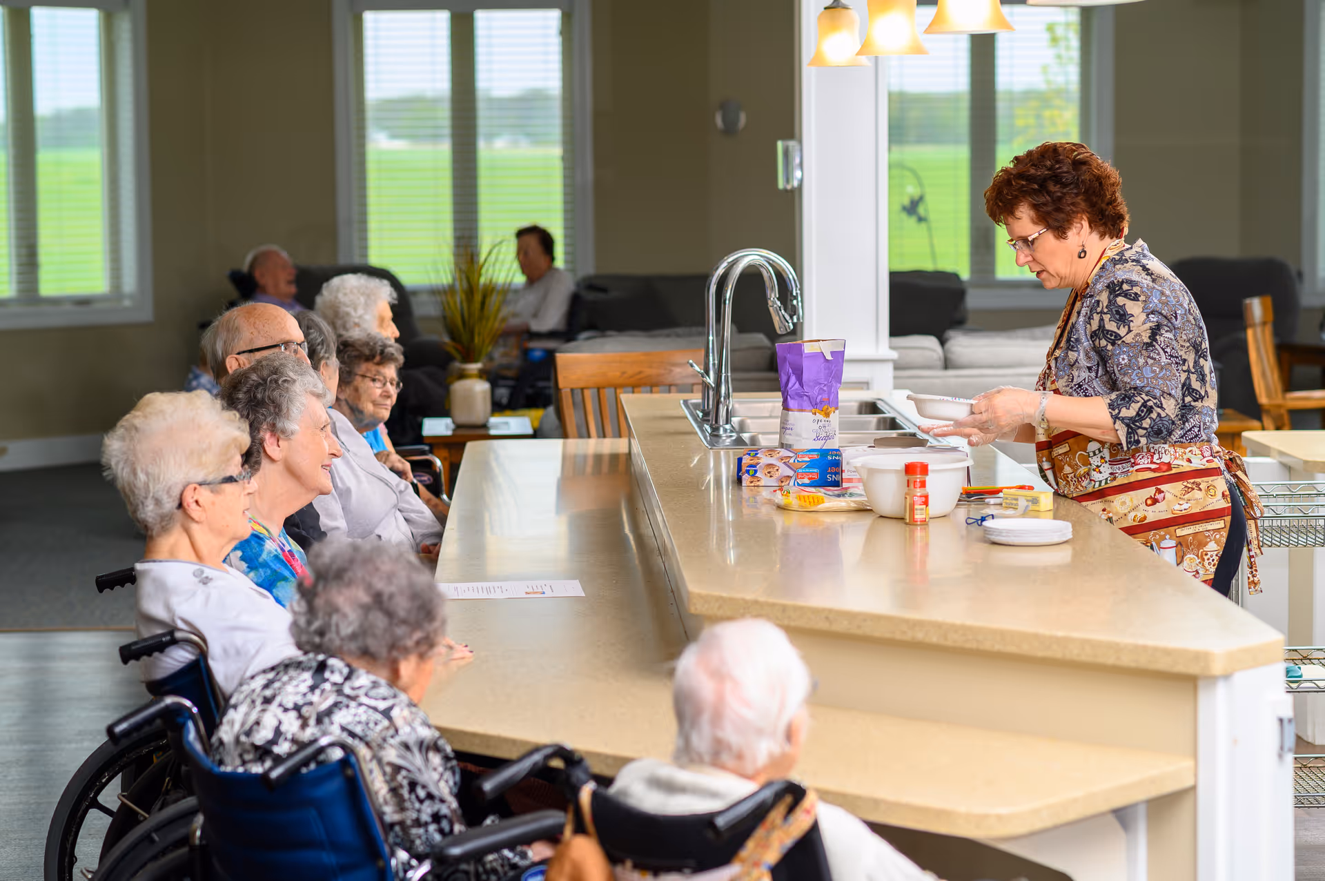 A group of elderly people seated at a long kitchen counter watching a woman in an apron preparing food. The setting is a bright room with large windows and comfortable seating in the background.