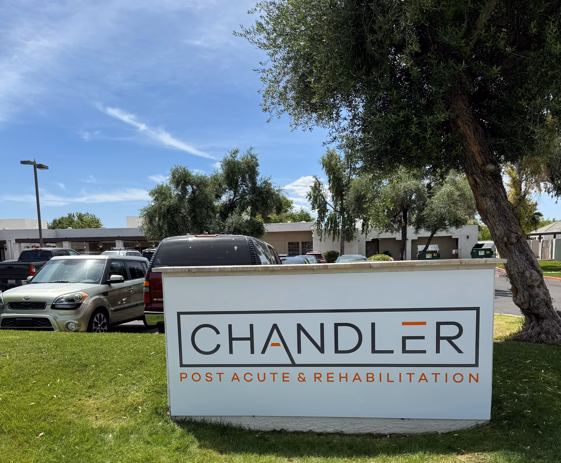 Outdoor view of a sign for Chandler Post Acute & Rehabilitation on a grassy area with trees and parked cars in the background under a partly cloudy sky.