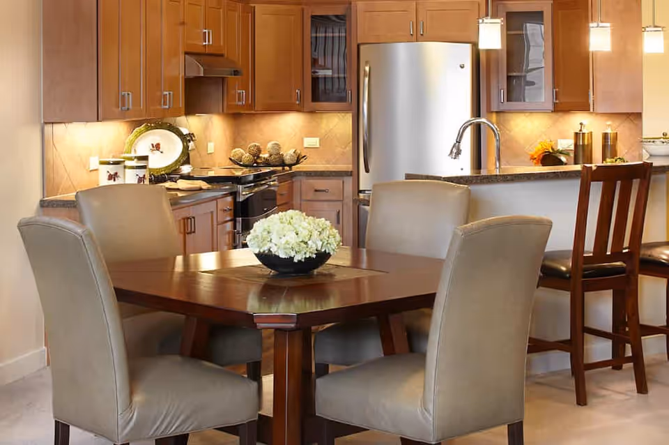 A modern kitchen and dining area with wooden cabinets, stainless steel refrigerator, stove, and a dining table with four beige upholstered chairs. A bowl of white flowers is placed on the dining table, and pendant lights hang above the kitchen counter with two wooden bar stools.