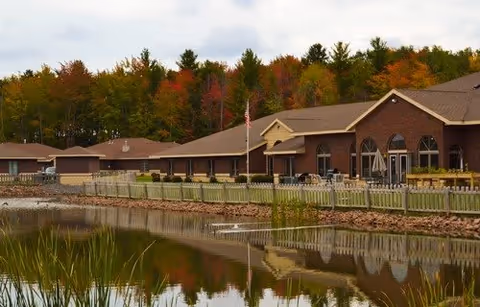 Exterior view of Willow Brooke Point Senior Living facility showing a long brick building with multiple windows and a flagpole in front. The building is adjacent to a calm pond with green reeds along the edge and trees with autumn foliage in the background.