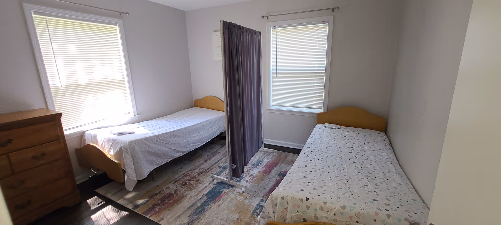 A small bedroom with two single beds separated by a privacy curtain on a stand. Each bed has a folded towel on top. There are two windows with blinds partially closed, a wooden dresser on the left side, and a colorful area rug on the floor.