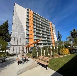 Exterior view of a multi-story apartment building with balconies, surrounded by trees and landscaped greenery. There are benches and a paved walkway in the foreground under a clear blue sky.
