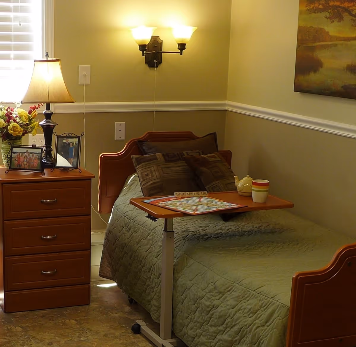A neatly made single bed in a senior living bedroom with an overbed table, nightstand, lamp, framed photos and wall sconce.