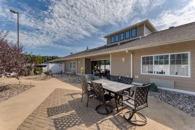Outdoor patio area at Cedarhurst of Breese featuring a table with six metal chairs on a paved surface, surrounded by landscaping with rocks and small bushes. The building exterior is beige with white trim and large windows, under a partly cloudy sky.