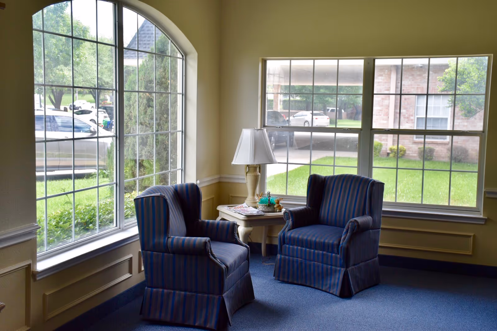 A cozy sitting area with two blue and red striped armchairs facing each other, separated by a small wooden side table with a white lamp and decorative items. Large windows behind the chairs provide a view of green grass, trees, and parked cars outside.