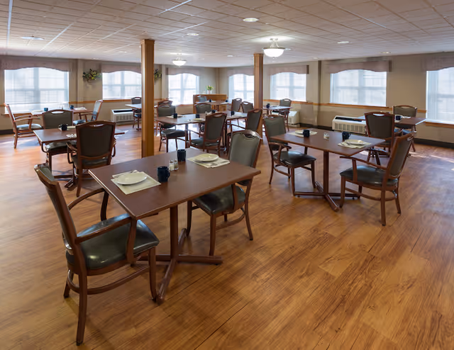 A spacious dining room with multiple wooden tables and chairs arranged neatly. Each table is set with placemats, plates, cups, and condiments. The room has large windows with blinds allowing natural light to fill the space, wooden flooring, and ceiling lights providing additional illumination.