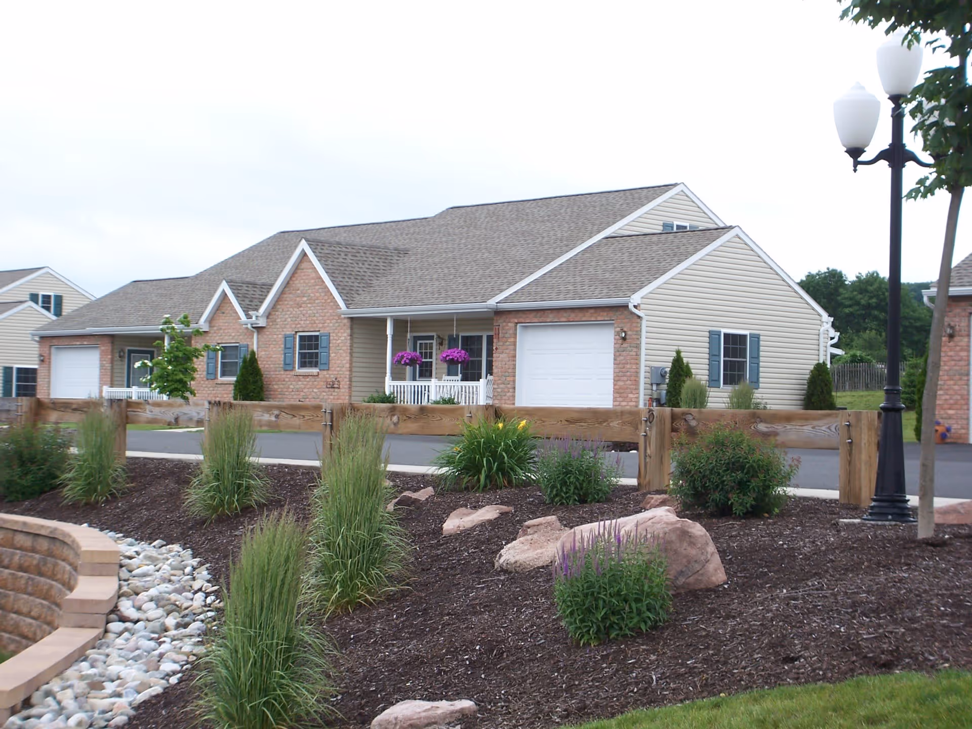 Exterior view of a single-story residential building with brick and siding facade, a garage, a small front porch with hanging flower baskets, and a landscaped garden with shrubs, rocks, and a lamp post in the foreground.