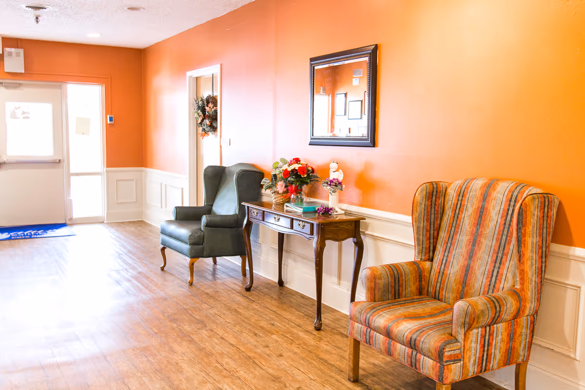 A hallway with orange walls and wood flooring featuring two armchairs, one striped and one solid dark green, separated by a wooden console table with flowers and decorative items. A mirror hangs on the wall above the table, and a door with a wreath is visible in the background near an exit door with a blue mat.