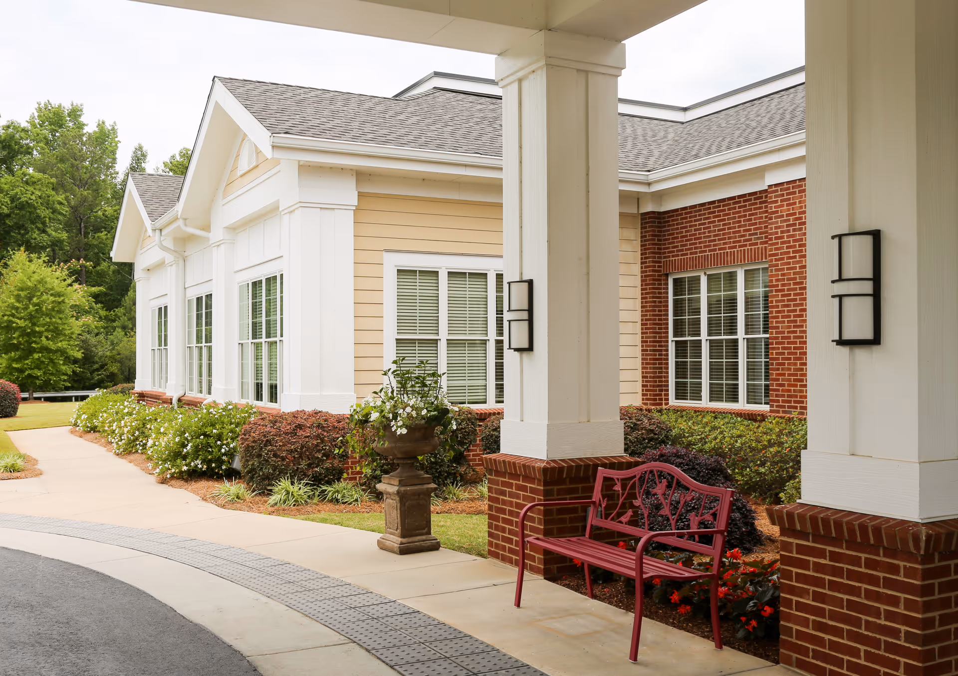 Exterior view of a senior living facility showing a covered entrance with white columns and brick bases. There is a red metal bench and a decorative planter with flowers near the entrance. The building has beige siding and red brick walls with multiple windows. Landscaping includes bushes and small trees along a curved sidewalk.