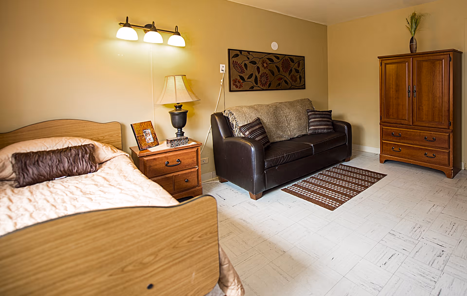 A cozy senior living bedroom with a bed, bedside table and lamp, a leather sofa, and a wooden armoire.