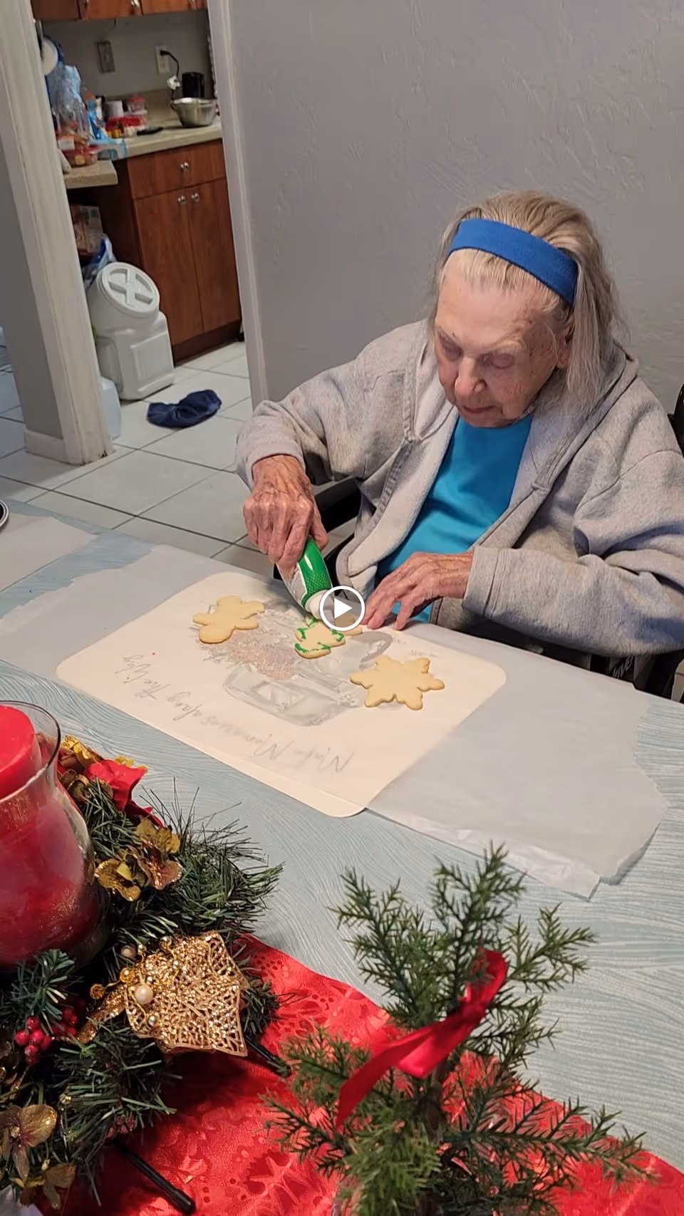 An elderly woman wearing a blue headband and gray sweater decorates Christmas cookies at a table. The table has a festive centerpiece with a red candle, greenery, and gold ornaments. The background shows a kitchen area with wooden cabinets and tiled floor.