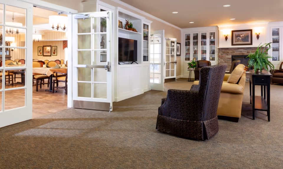 A cozy living room area with patterned armchairs, a beige sofa, a fireplace with stone surround, and built-in bookshelves. To the left, glass double doors open to a dining room with tables and chairs, softly lit by chandeliers.